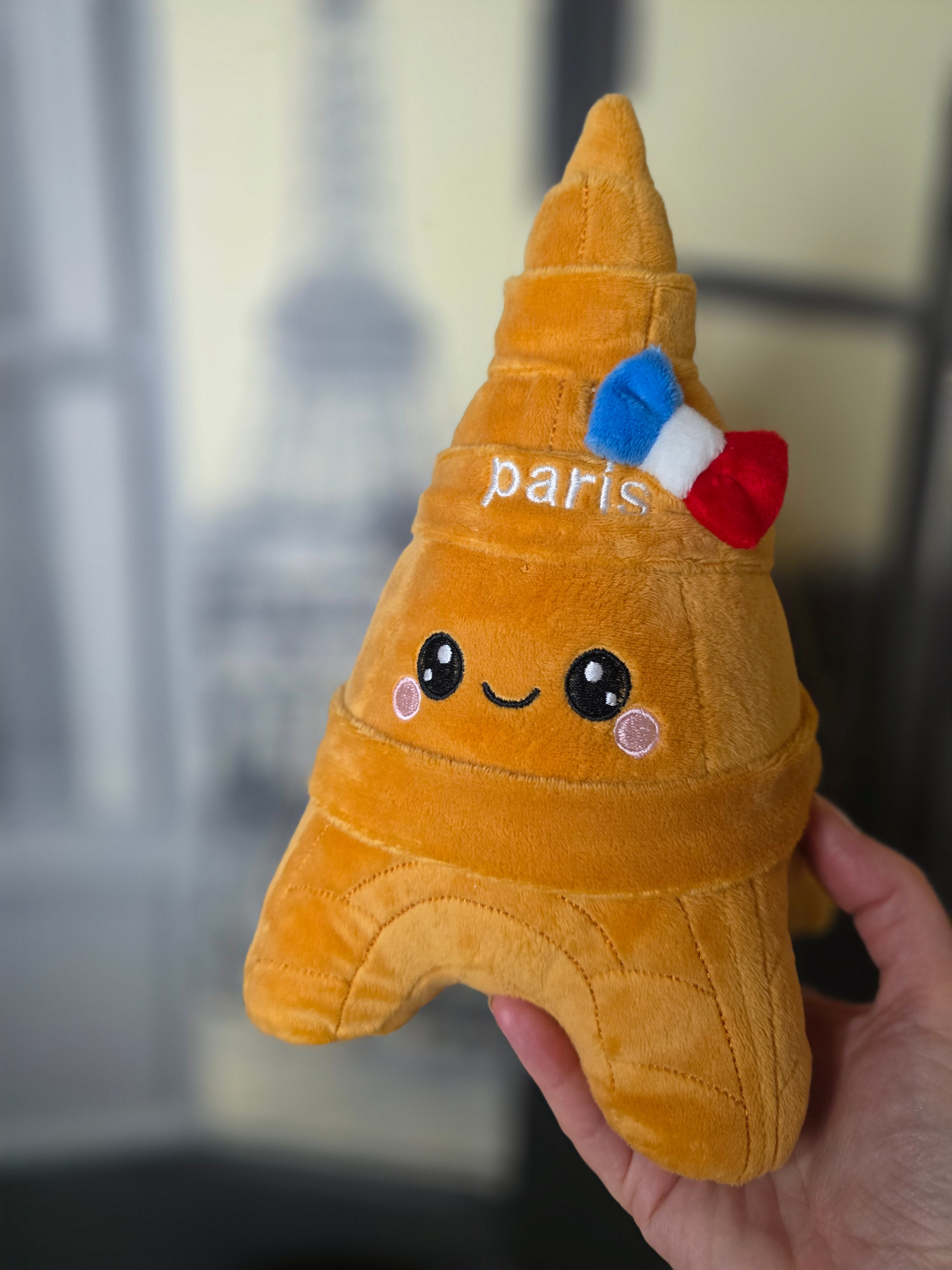 A plush toy shaped like the Eiffel Tower in golden-brown fabric. The plush has embroidered facial features, a small red, white, and blue bow, and the word 'paris' stitched on the front.